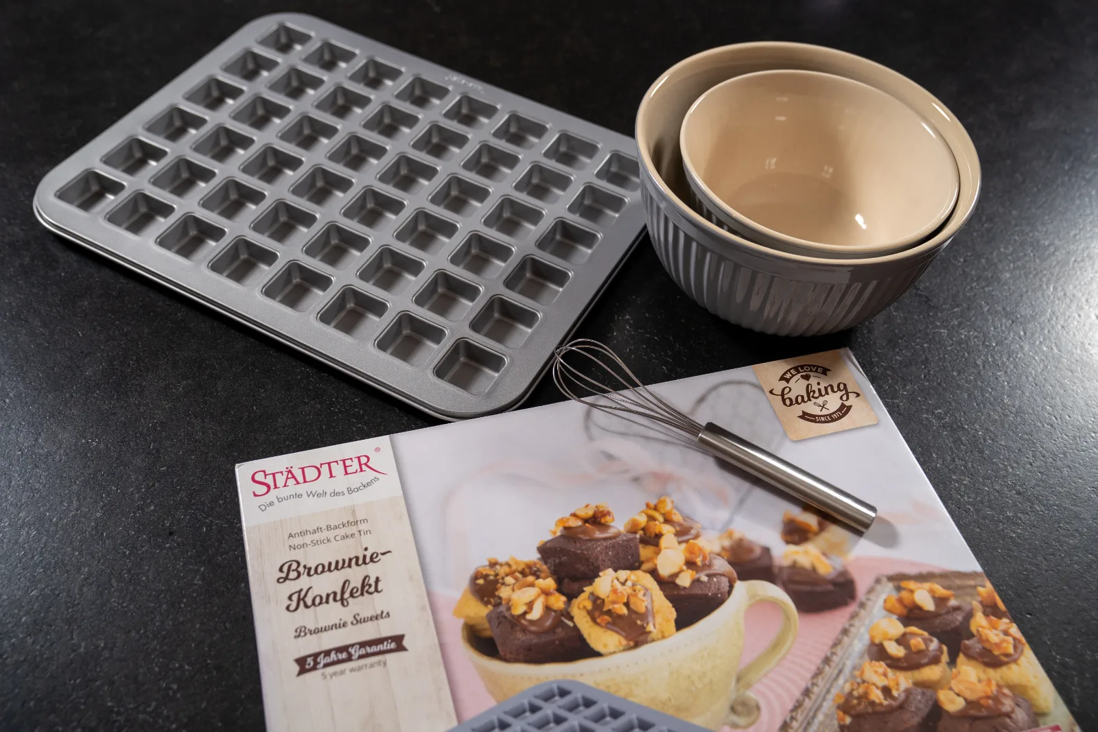 Brownie confectionery non-stick baking tin with mixing bowls and whisk on a dark work surface