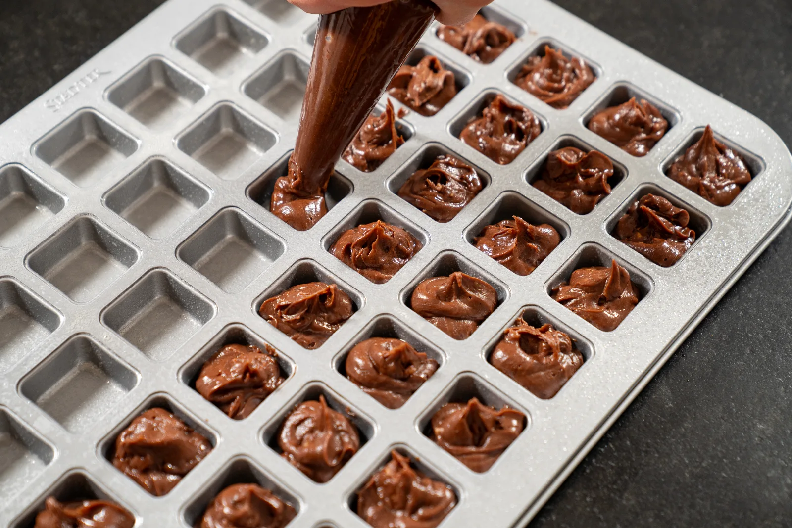 Brownie dough being piped into the cavities of the baking tin