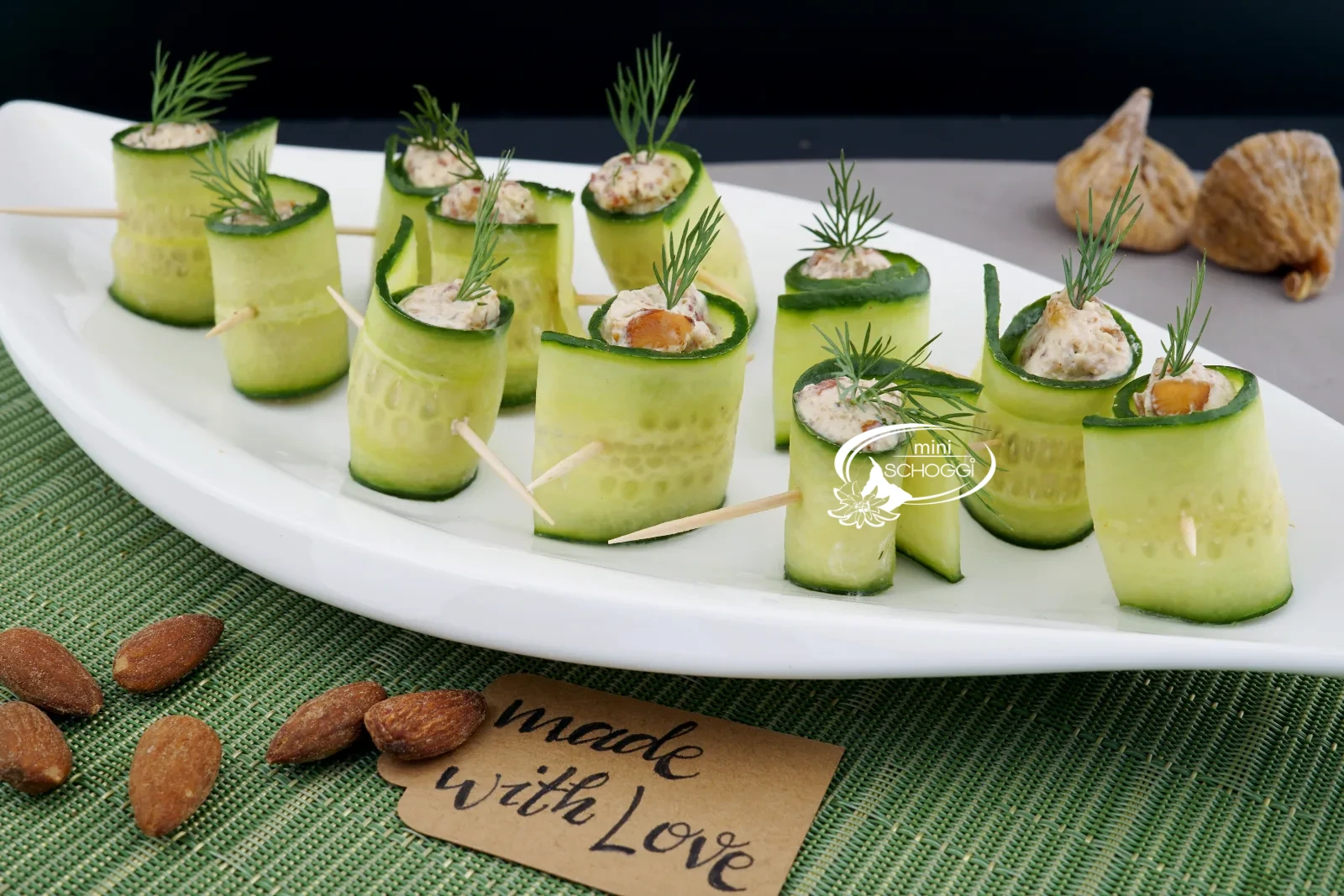 Involtini di cetriolo ripieni con aneto su un piatto bianco, sullo sfondo due bicchieri di vino