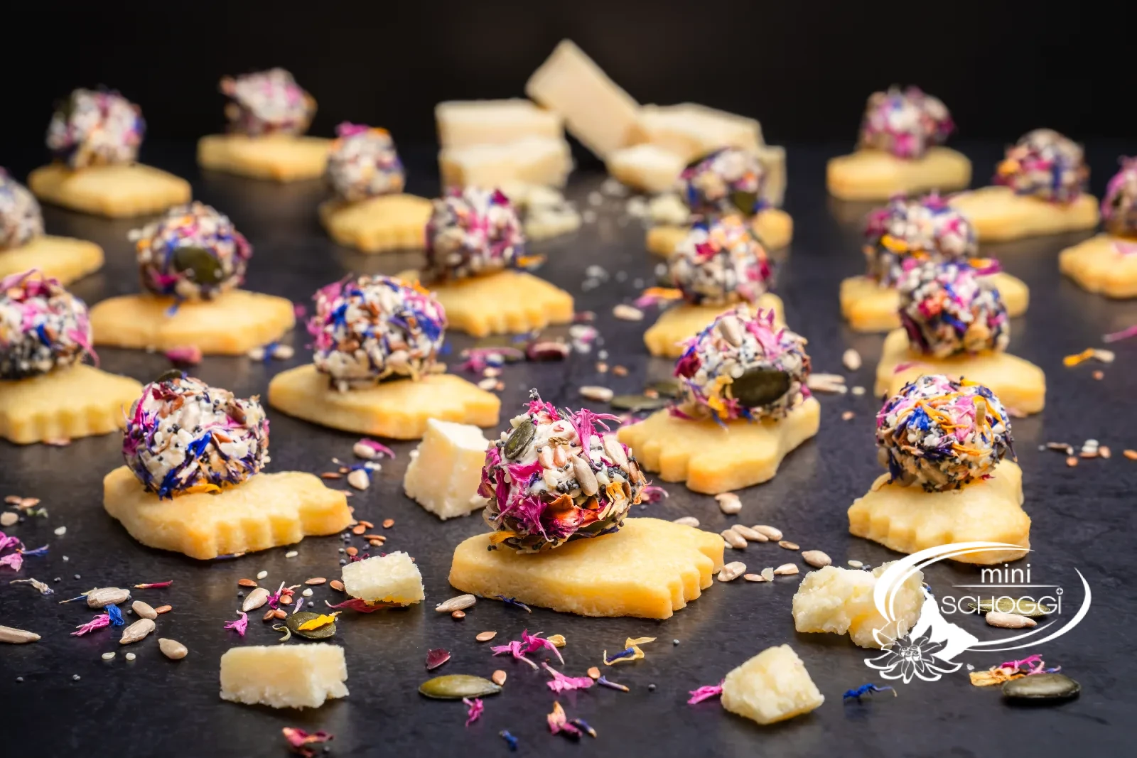 Piedi puzzolenti da aperitivo con palline colorate di formaggio fresco, accanto pezzi di parmigiano e un bicchiere con la miscela di fiori e semi