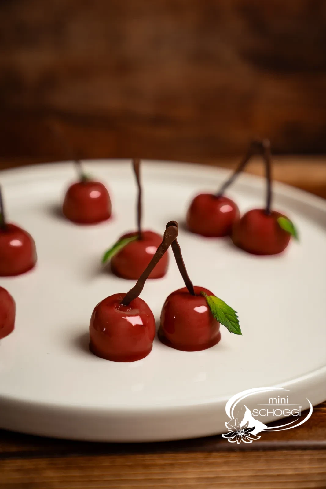 Mehrere glänzende Mousse-Kirschen mit Schokoladenstielen und Minzblättern auf einer hellen Platte, täuschend echt wirkend