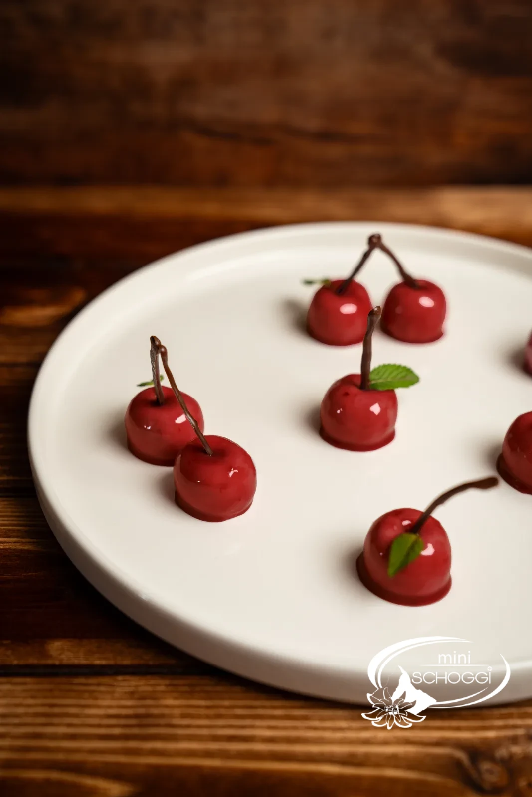 Mehrere glänzende Mousse-Kirschen mit Schokoladenstielen und Minzblättern auf einer hellen Platte, täuschend echt wirkend