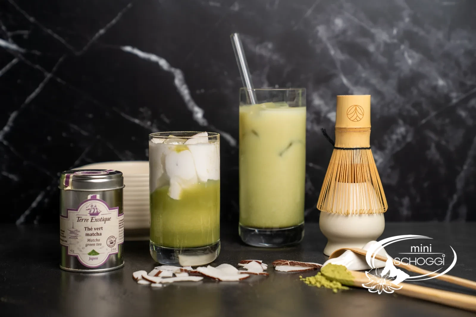 Two glasses of matcha iced latte with a bamboo whisk and matcha tin on a light surface