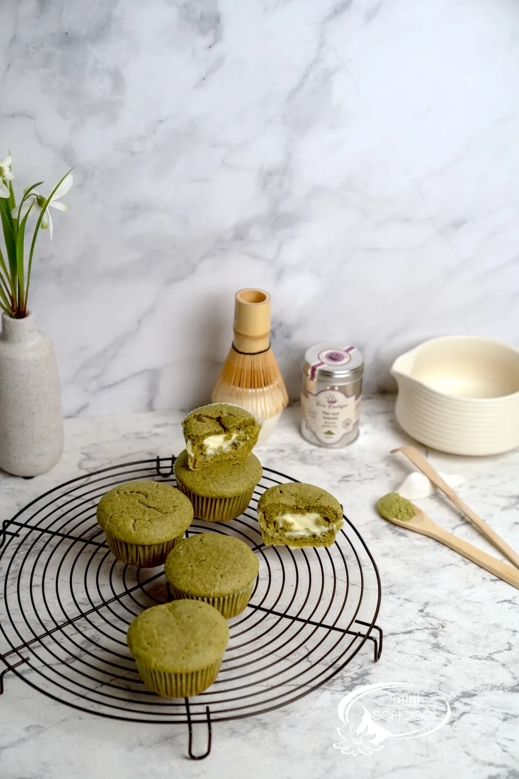Matcha Muffins mit cremigem Frischkäsekern auf dem Kuchengitter, dazu Matcha-Dose und Bambusbesen