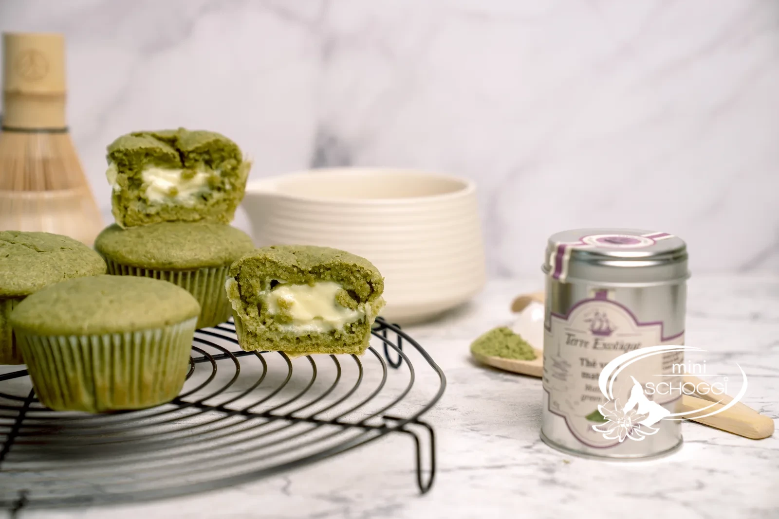 Matcha Muffins mit cremigem Frischkäsekern auf dem Kuchengitter, dazu Matcha-Dose und Bambusbesen